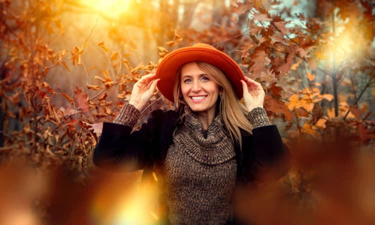 Middle-aged woman with dental implants smiles as she wears a red hat and sweater by trees with fall leaves
